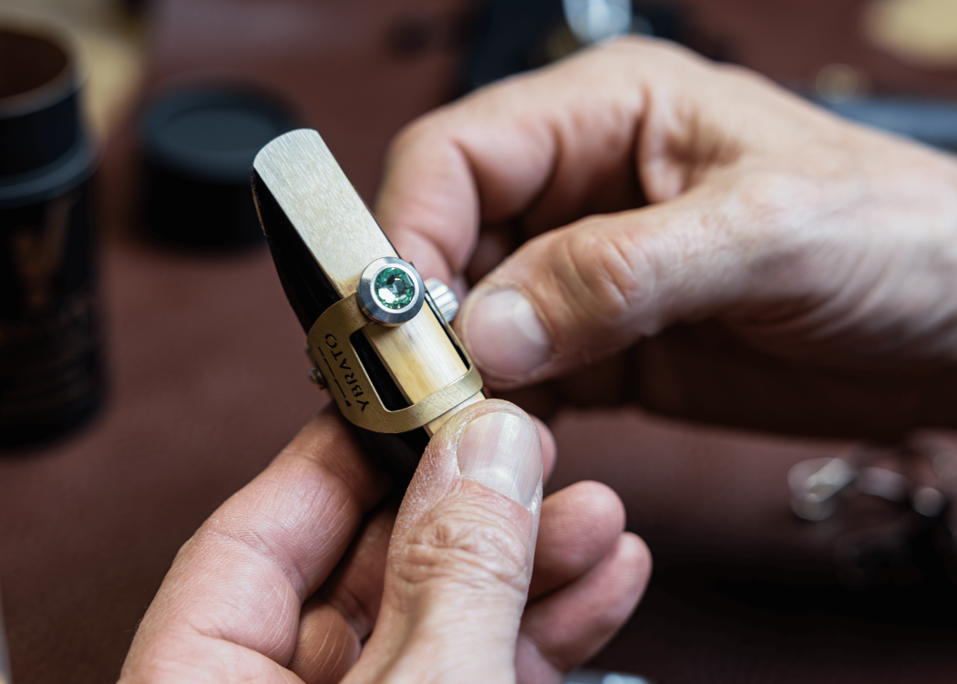 Close-up of a craftsman's hands assembling a YBRATO ReedReflector ligature in the Crystal Edition. The image shows the brass ligature mounted on a mouthpiece with a vibrant green Swarovski crystal being meticulously fitted.