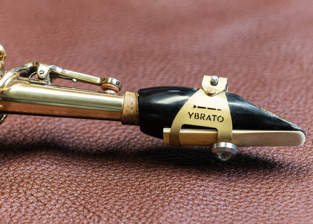 Side view of the YBRATO ReedReflector mounted on a saxophone mouthpiece. The image shows the gold-finished ligature with its engraved logo on the underside, resting on a brown leather surface.