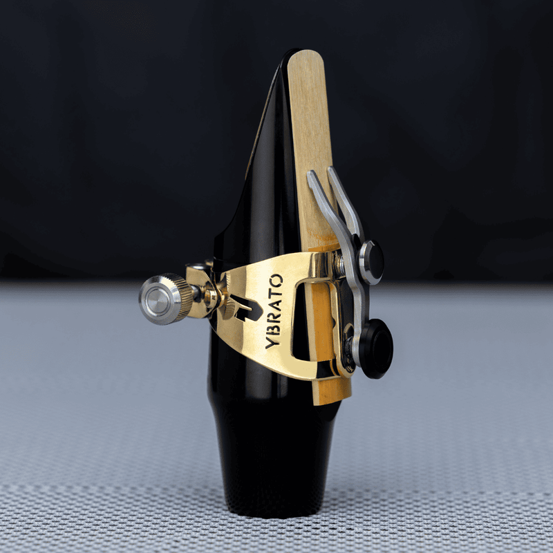 A close-up of the modern YBRATO SweetSpot Ligature in a polished gold finish, mounted on a black saxophone mouthpiece. The image showcases the high-precision CNC-milled design and the innovative adjustable mechanism for optimized reed vibration.